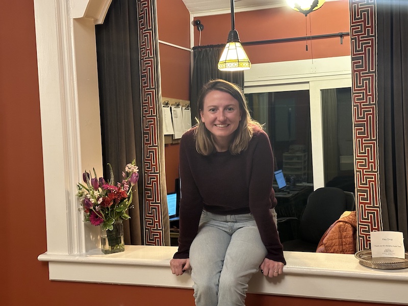 Claire Collery, proprietor of the inn, sitting by the roaring fireplace in the main lobby in a maroon sweater and blue jeans, legs crossed. She is inexplicably holding a newspaper open over her head, creating a sort of roof shape.
